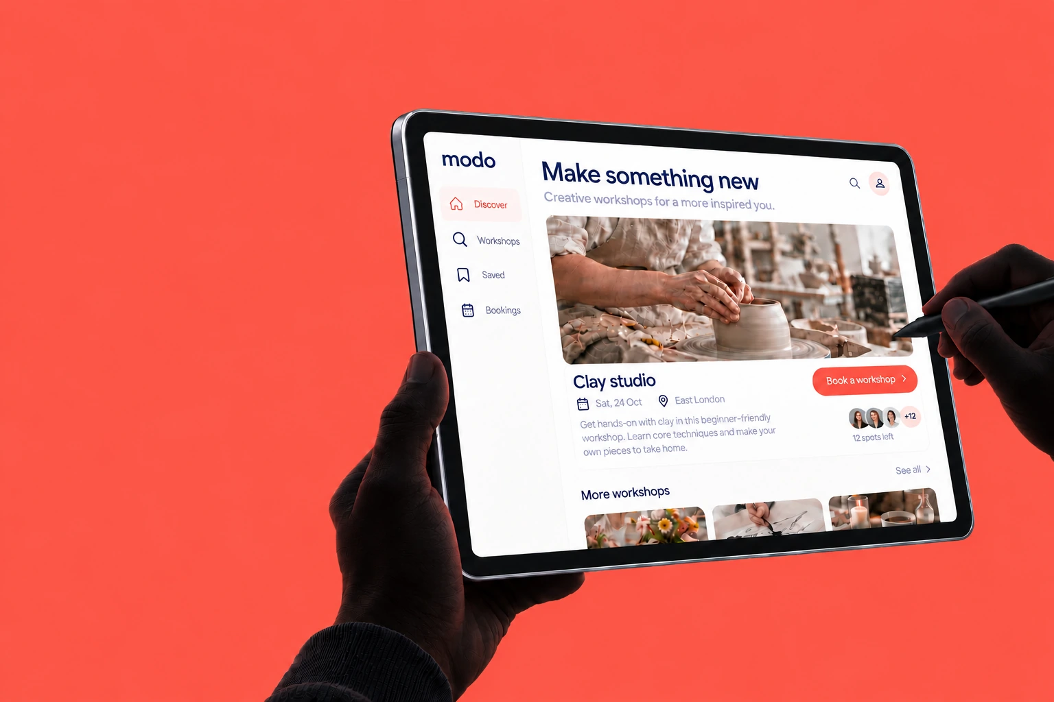 A shadowed left hand holding an iPad with the Modo interface, with a right hand holding a pen against a coral background